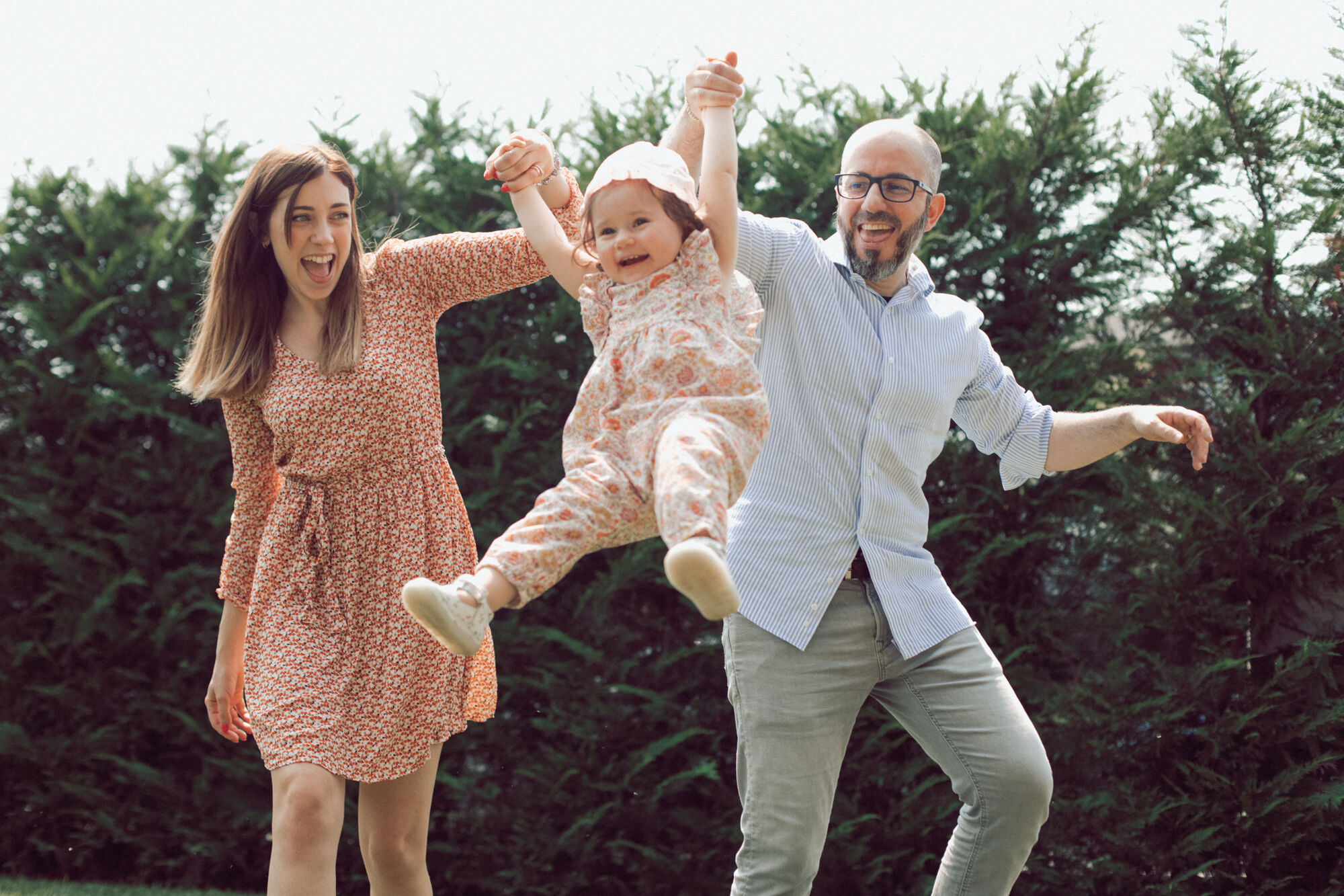 Genitori fanno volare in alto la figlia ridendo davanti alla siepe, servizio fotografico famiglia in giardino