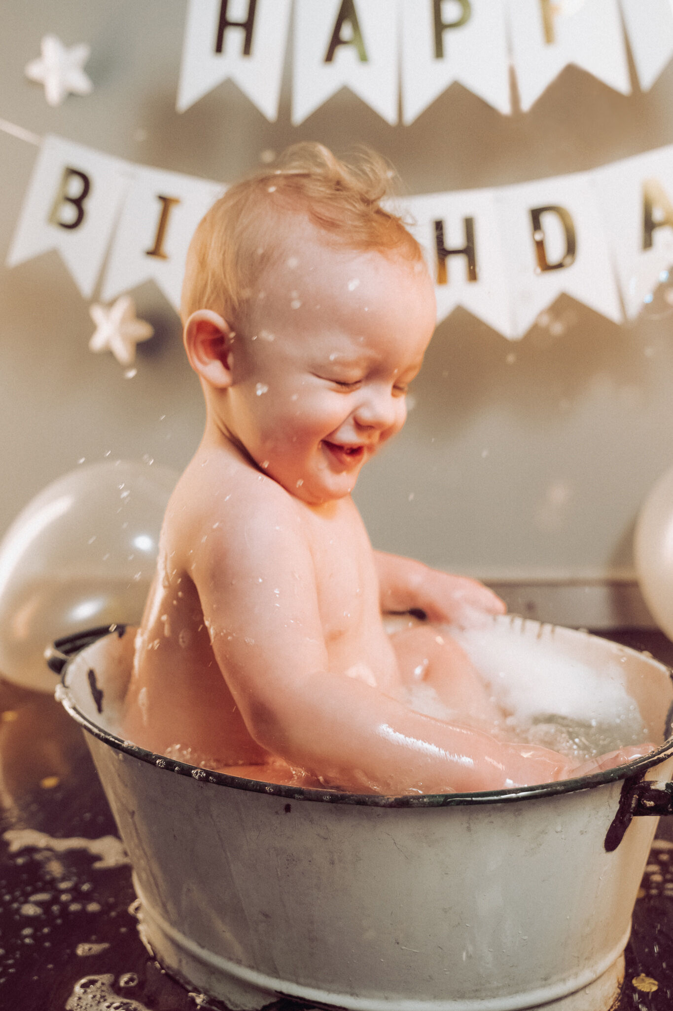 Bimbo sorridente schizza acqua nella tinozza di smalto sotto il festone Happy Birthday, dopo lo smash cake