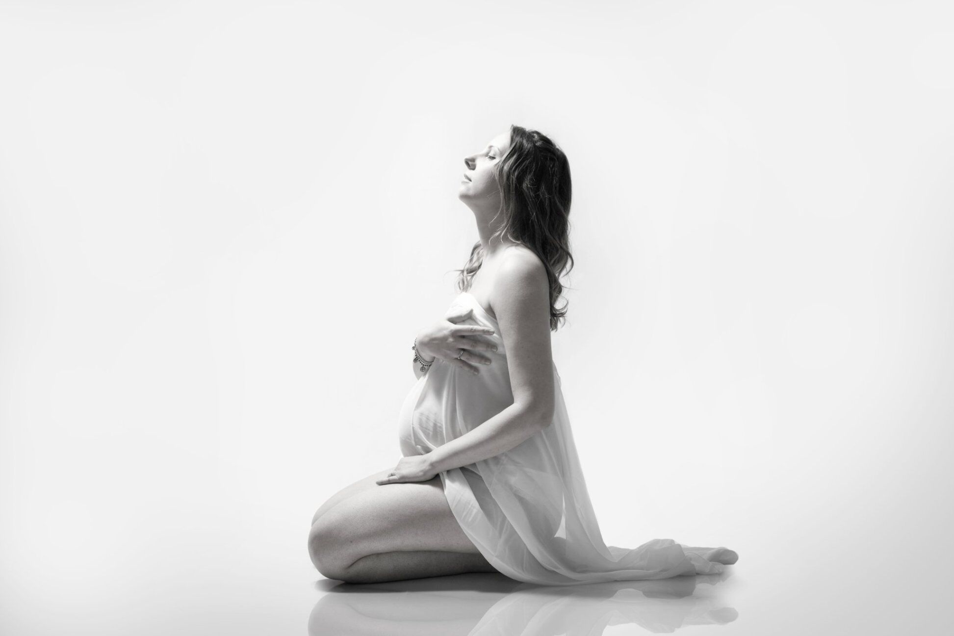 Futura mamma inginocchiata avvolta in un velo bianco, viso verso la luce, servizio fotografico gravidanza in bianco e nero