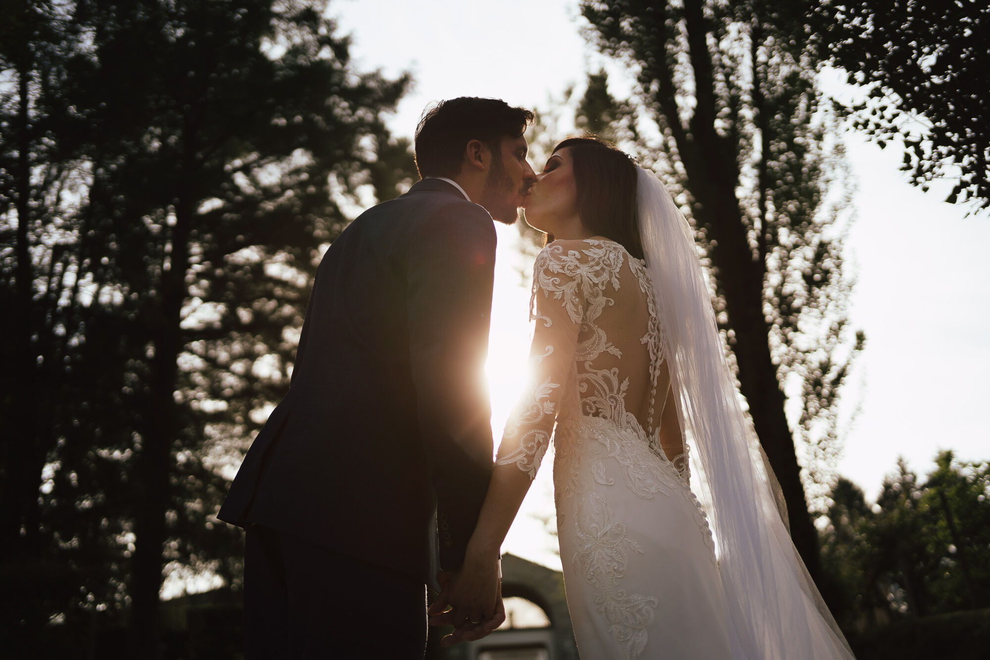 Bacio degli sposi in controluce al tramonto, sole tra le figure e alberi scuri